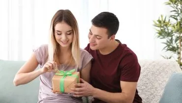 Imagem mostra um casal apaixonado. Ele está dando um presente para ela em uma caixa bege com laço verde.