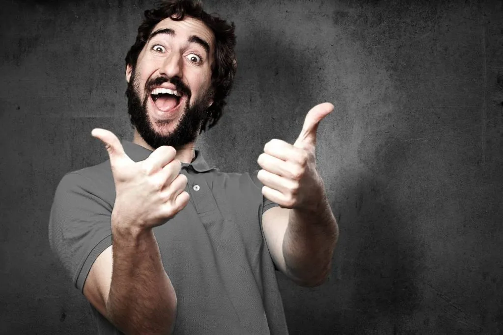 Imagem mostra um homem rindo e fazendo joinha com as duas mãos. Ele está usando uma camisa polo cinza em um fundo cinza.