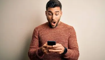 Imagem mostra um homem assustado olhando o celular. Ele tem barba e usa uma blusa de cor bege escuro