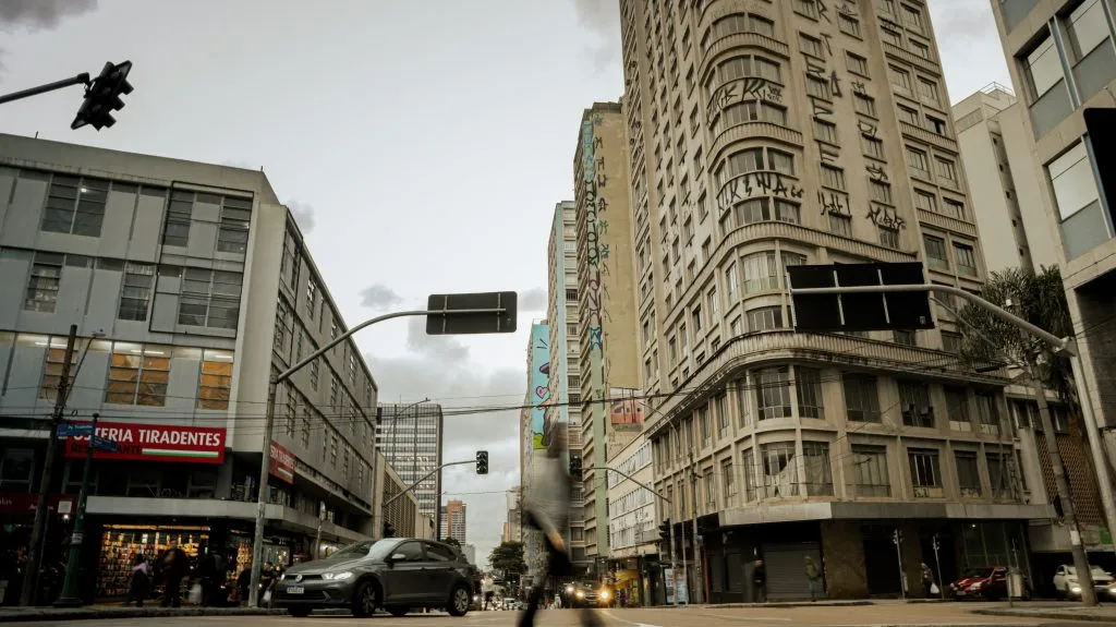 Fotografia em plano geral e ângulo contra-plongée (de baixo para cima) de uma rua urbana movimentada entre edifícios altos, captada ao entardecer na região central de Curitiba. Em primeiro plano, o asfalto da rua exibe pedestres e veículos em circulação, com destaque para um carro cinza escuro trafegando em direção ao lado esquerdo e a silhueta em movimento acelerado (efeito motion blur) de um transeunte no centro inferior. Placas de sinalização viária suspensas por hastes metálicas curvas projetam-se sobre a via. À esquerda, o piso térreo de um edifício comercial mostra o letreiro vermelho com letras brancas de uma "RESTAURANTE / OSTERIA TIRADENTES", além de vitrines iluminadas. No plano intermediário, imponentes edifícios comerciais e residenciais com fachadas de janelas repetitivas flanqueiam a via. O edifício à direita destaca-se por sua fachada curva adornada com pichações e graffitis nos andares superiores. Ao fundo, a perspectiva da rua estende-se em direção a outros prédios e árvores sob um céu nublado com iluminação difusa de fim de tarde.