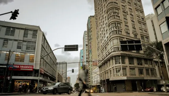 Fotografia em plano geral e ângulo contra-plongée (de baixo para cima) de uma rua urbana movimentada entre edifícios altos, captada ao entardecer na região central de Curitiba. Em primeiro plano, o asfalto da rua exibe pedestres e veículos em circulação, com destaque para um carro cinza escuro trafegando em direção ao lado esquerdo e a silhueta em movimento acelerado (efeito motion blur) de um transeunte no centro inferior. Placas de sinalização viária suspensas por hastes metálicas curvas projetam-se sobre a via. À esquerda, o piso térreo de um edifício comercial mostra o letreiro vermelho com letras brancas de uma "RESTAURANTE / OSTERIA TIRADENTES", além de vitrines iluminadas. No plano intermediário, imponentes edifícios comerciais e residenciais com fachadas de janelas repetitivas flanqueiam a via. O edifício à direita destaca-se por sua fachada curva adornada com pichações e graffitis nos andares superiores. Ao fundo, a perspectiva da rua estende-se em direção a outros prédios e árvores sob um céu nublado com iluminação difusa de fim de tarde.