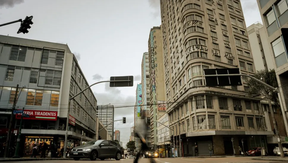Fotografia em plano geral e ângulo contra-plongée (de baixo para cima) de uma rua urbana movimentada entre edifícios altos, captada ao entardecer na região central de Curitiba. Em primeiro plano, o asfalto da rua exibe pedestres e veículos em circulação, com destaque para um carro cinza escuro trafegando em direção ao lado esquerdo e a silhueta em movimento acelerado (efeito motion blur) de um transeunte no centro inferior. Placas de sinalização viária suspensas por hastes metálicas curvas projetam-se sobre a via. À esquerda, o piso térreo de um edifício comercial mostra o letreiro vermelho com letras brancas de uma "RESTAURANTE / OSTERIA TIRADENTES", além de vitrines iluminadas. No plano intermediário, imponentes edifícios comerciais e residenciais com fachadas de janelas repetitivas flanqueiam a via. O edifício à direita destaca-se por sua fachada curva adornada com pichações e graffitis nos andares superiores. Ao fundo, a perspectiva da rua estende-se em direção a outros prédios e árvores sob um céu nublado com iluminação difusa de fim de tarde.