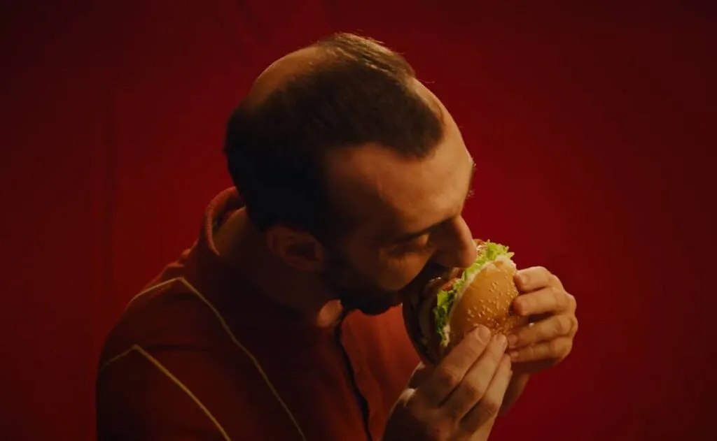 Imagem mostra um homem calvo comento um sanduíche no drive thru do Burguer King