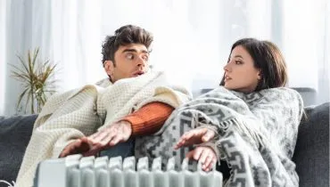 A foto mostra um homem e uma mulher agasalhados com cobertas, sentados em um sofá, com as mãos esticadas em cima de um aquecedor elétrico.