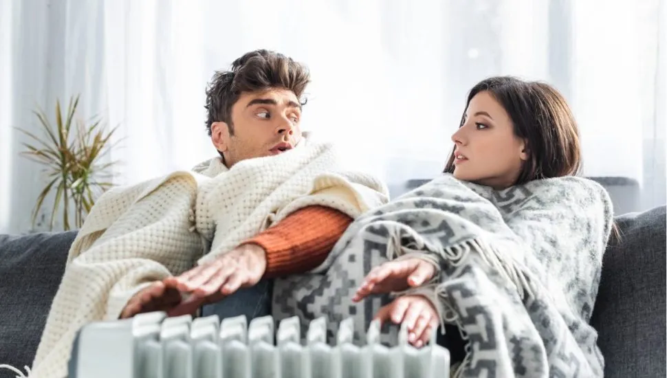 A foto mostra um homem e uma mulher agasalhados com cobertas, sentados em um sofá, com as mãos esticadas em cima de um aquecedor elétrico.