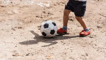 Imagem mostra um meino, uma criança, chutando uma bola num piso de terra. parece bem humilde