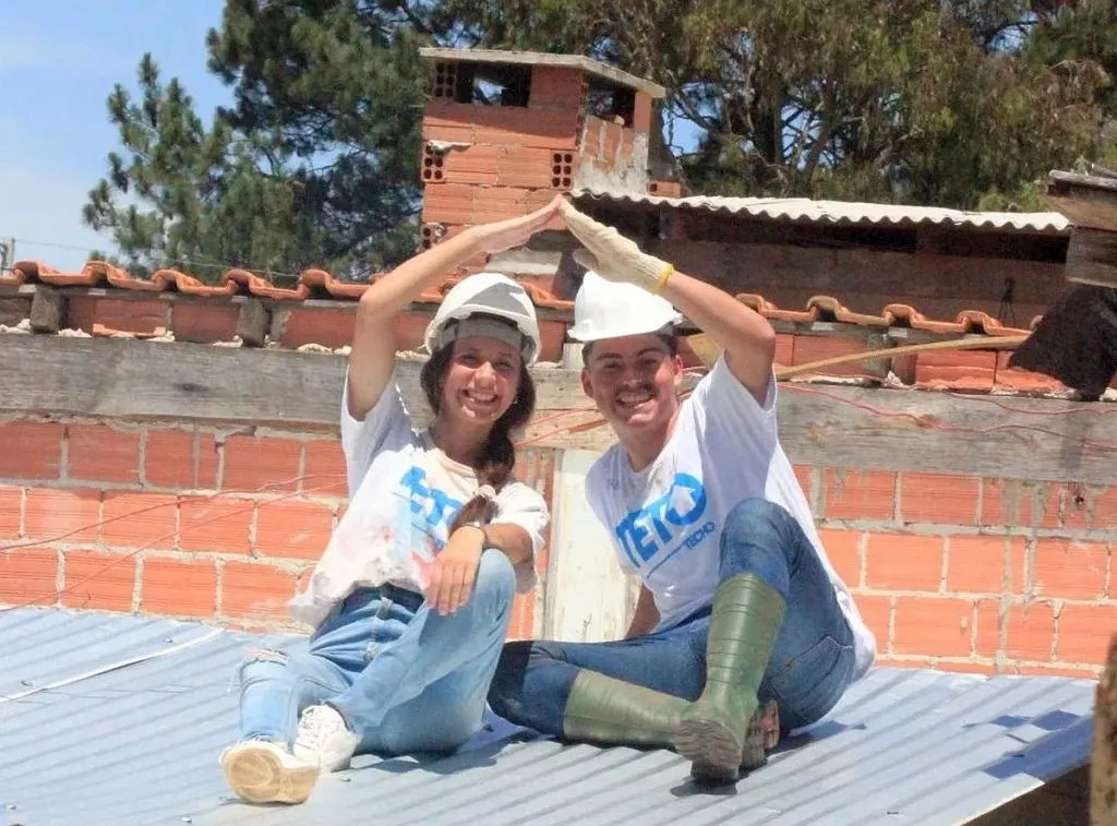 Imagme mostra um homem e uma mulher, emcimade um telhado de uma casa humilde, juntando as mãos para formar o desenho de um teto