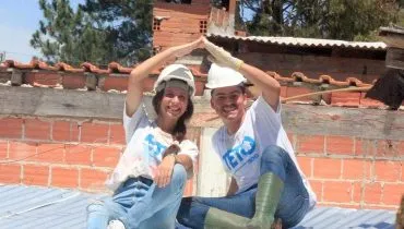 Imagme mostra um homem e uma mulher, emcimade um telhado de uma casa humilde, juntando as mãos para formar o desenho de um teto