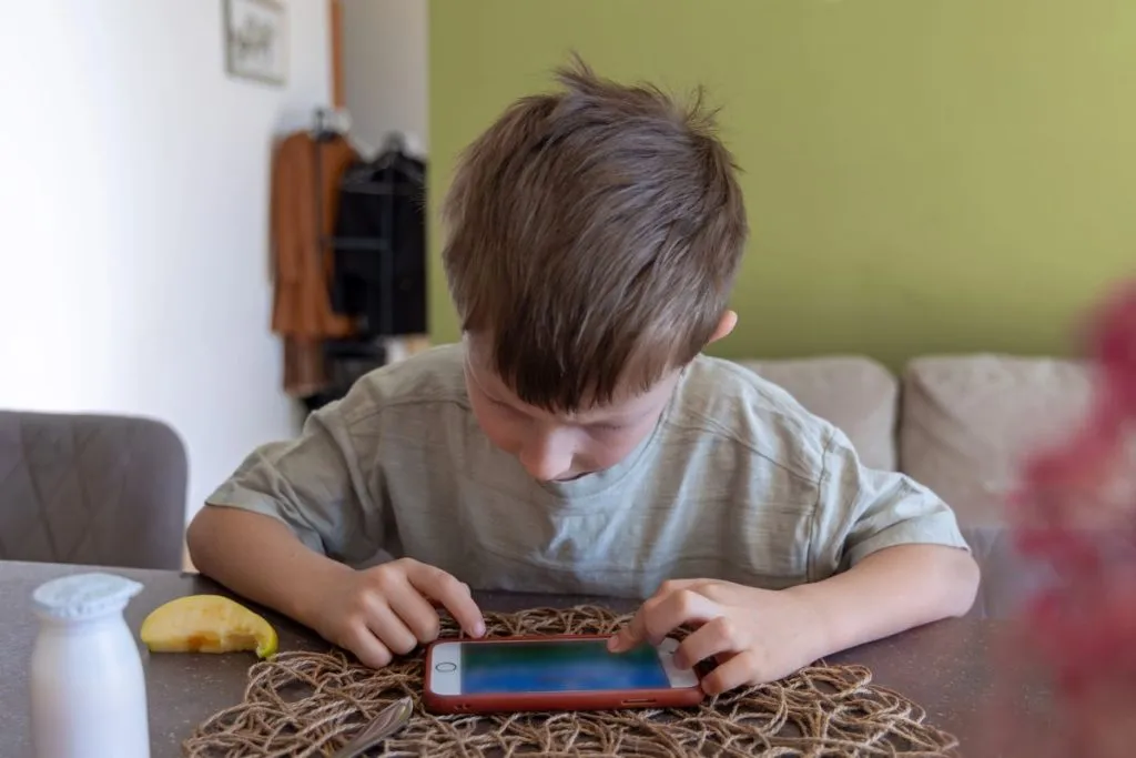 na imagem uma criança está vidrada num celular, que está sobre a mesa