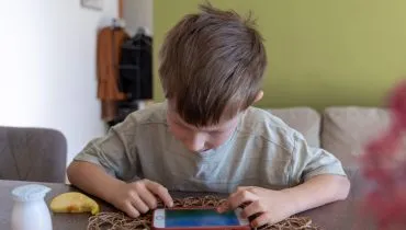 na imagem uma criança está vidrada num celular, que está sobre a mesa