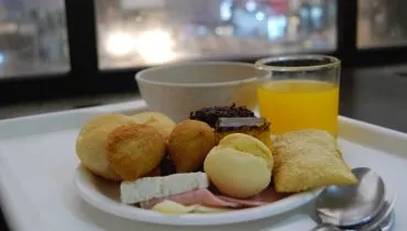 Prato pequeno com coxinha, risoles, pão de queijo, frios, pão, bolo de chocolate, bolo de cenoura, pastel. Copo com suco. Cumbuca com sopa.